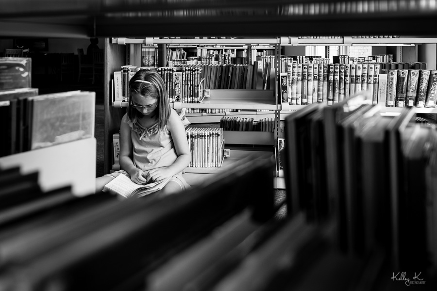 Black and white image of girl reading in a library through the stacks